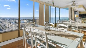 A high-rise room with a large glass table and chairs, a view of the city and ocean, and a ceiling fan. - Capri 7: 2 BR, 2 BA Condominium in Ocean City, Sleeps 6 (Ocean City)
