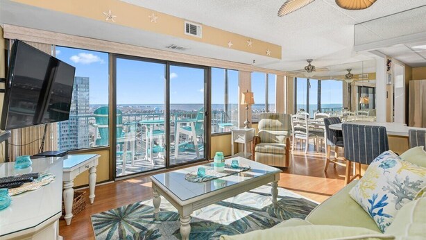 A spacious living room with large windows, a flat-screen TV, a dining area, and a balcony with a view of the city. - Capri 7: 2 BR, 2 BA Condominium in Ocean City, Sleeps 6 (Ocean City)