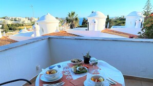 Une table dressée pour deux personnes sur un balcon, offrant une vue dégagée sur la mer et les bâtiments blancs en arrière-plan.
