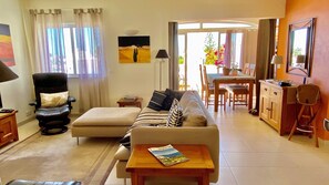 A living room with a sofa, armchair, dining table, and chairs. There is a lamp, a magazine on a table, and a painting on the wall. - Casa Papaya (Carvoeiro)