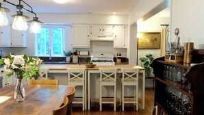 A modern kitchen with white cabinetry, a wooden dining table, and a bookshelf filled with books. - Cheerful 3-bedroom cottage with woodstove (Sheffield)