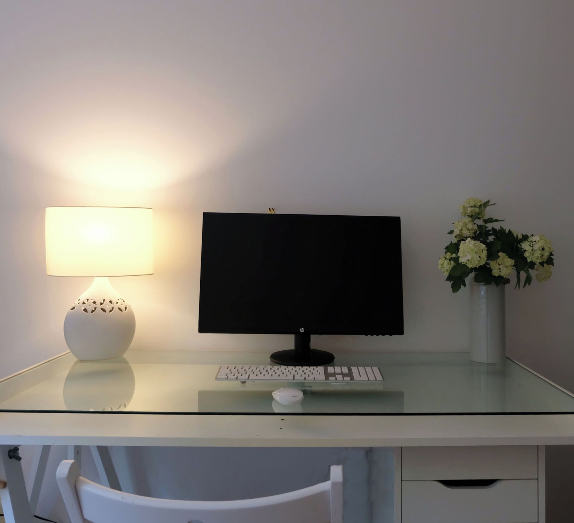 A desk with a computer monitor, keyboard, and mouse. There is a lamp and a vase with flowers on the desk.