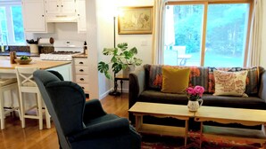 A living room with a dark blue armchair, a wooden coffee table, and a sofa with patterned cushions. A kitchen is visible in the background with white cabinets and a stove. - Cheerful 3-bedroom cottage with woodstove (Sheffield)