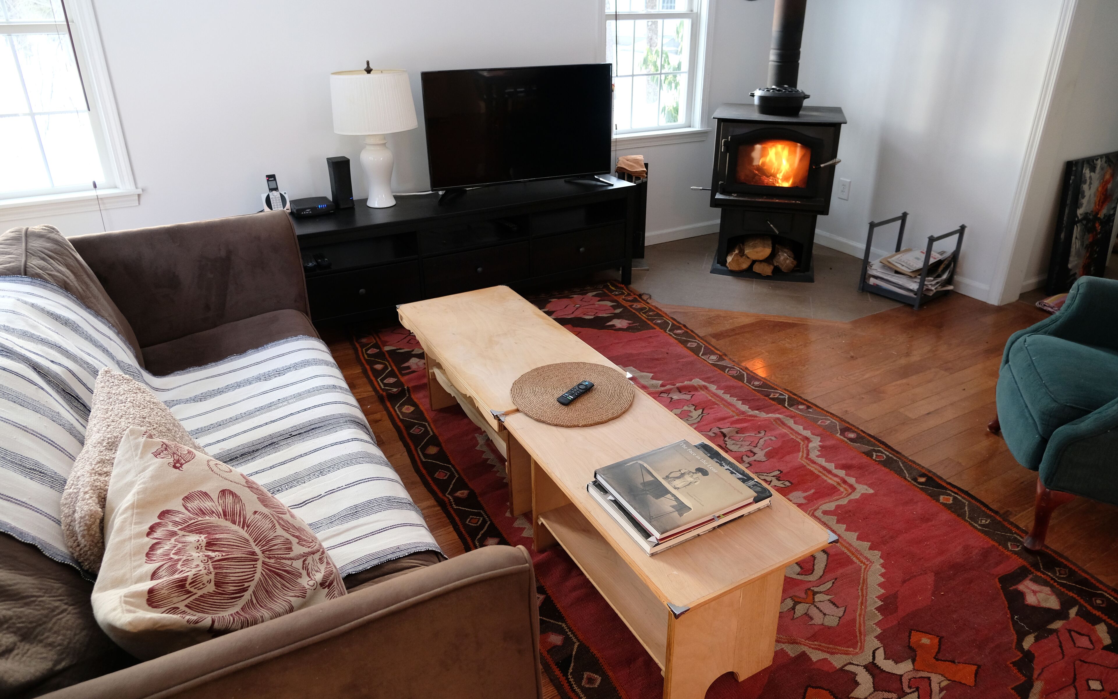 A cozy living room with a sofa, a wooden coffee table with a book and remote, a TV, a lamp, and a wood-burning stove.