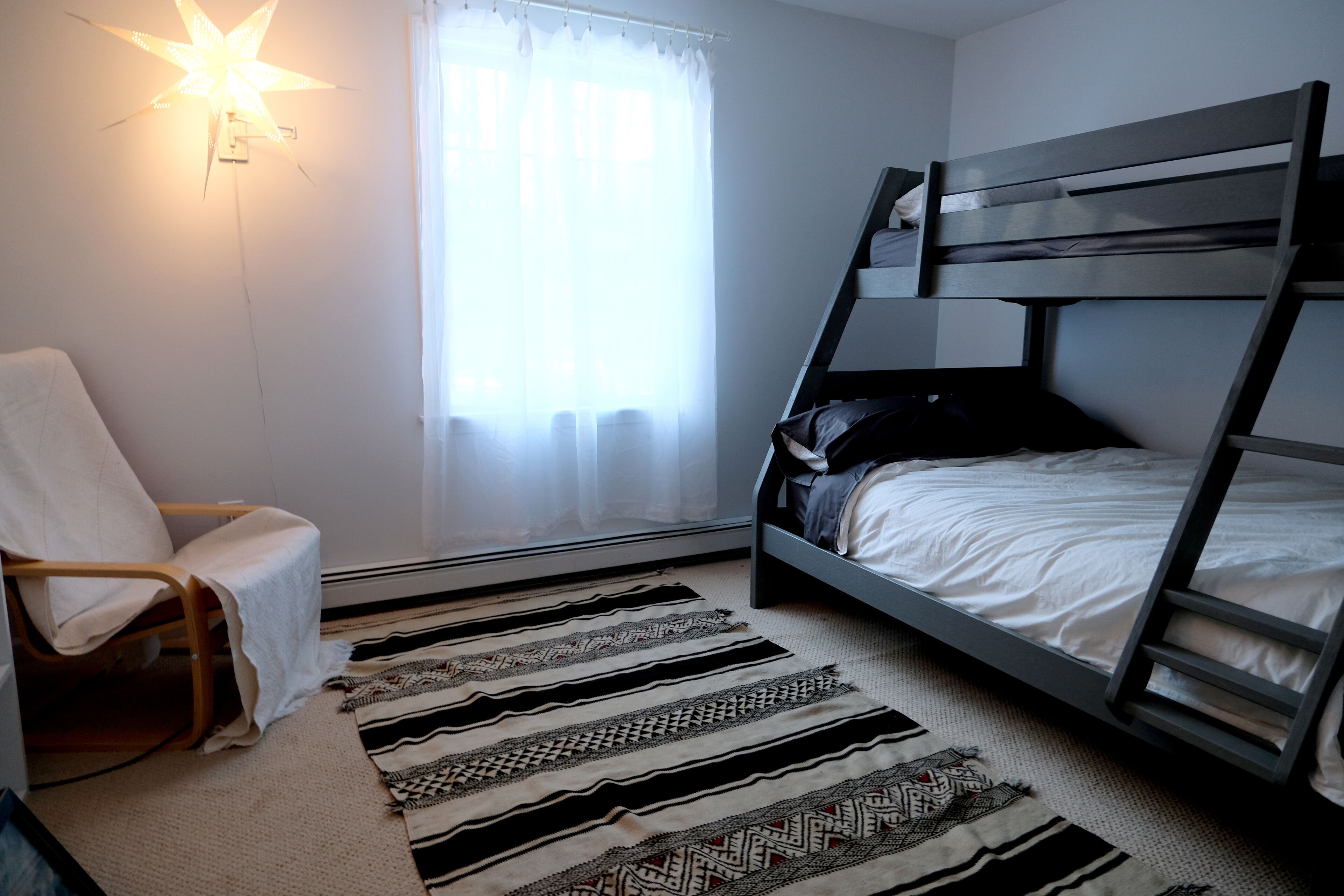Een kamer met een stapelbed, een stoel, een wandlamp in de vorm van een ster en een raam met dunne gordijnen.
