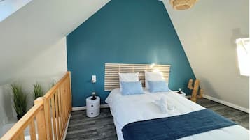 Een slaapkamer met een schuine balkenlaag, een houten hoofdbord, een bed met wit beddengoed, een blauwe accentmuur, een houten leuning en een kleine plant.