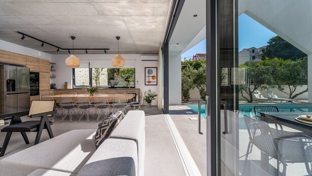 Cocina moderna con encimera de madera y electrodomésticos de acero inoxidable, zona de comedor con una mesa larga y sillas, y vistas a una piscina y vegetación exterior.