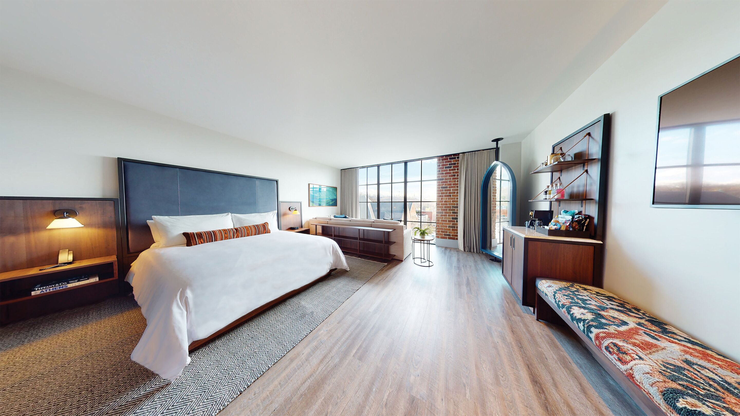Habitación de hotel moderna con una cama grande, un sofá, un escritorio y vistas a la ciudad a través de amplias ventanas.