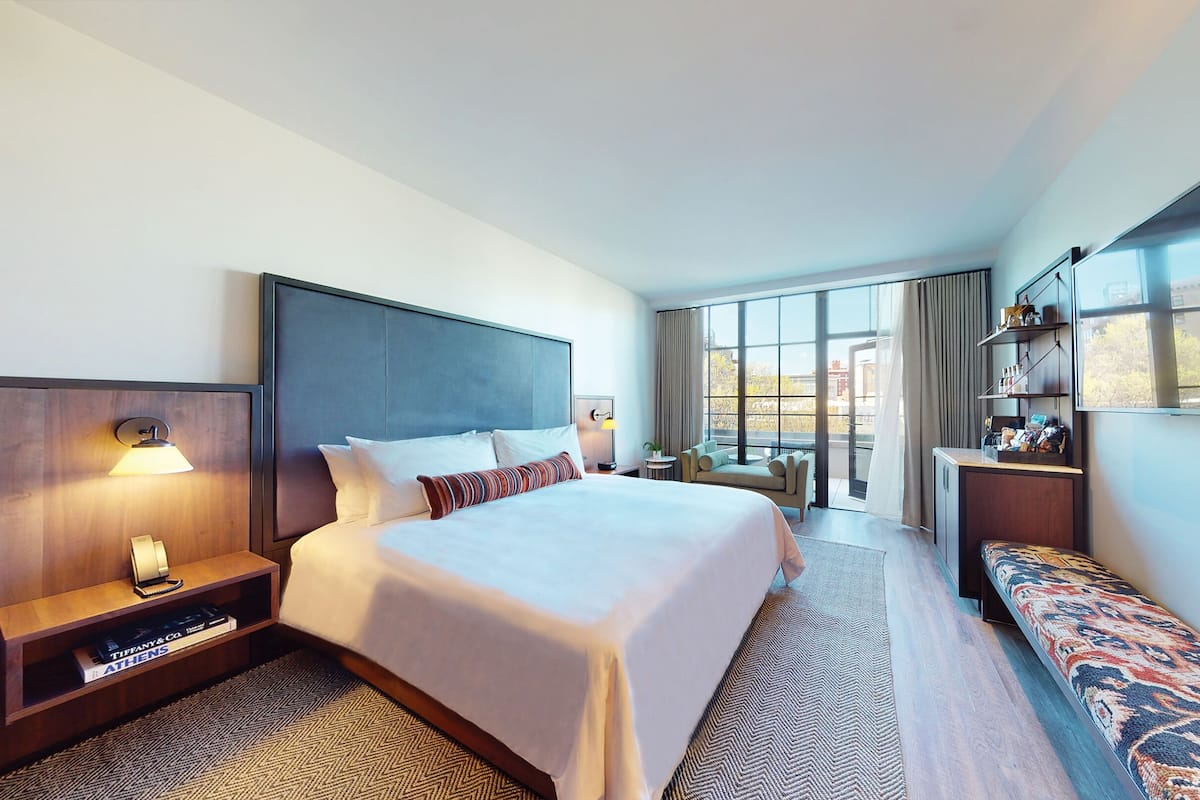 A hotel room with a large bed, a bedside table with a lamp, a bench, a small desk with shelves, and a large window offering a city view.