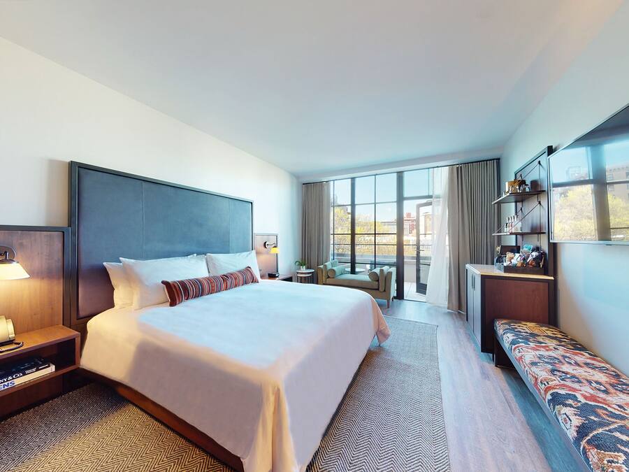 Quarto de hotel com uma cama grande, uma mesinha de cabeceira com abajur, um banco, uma pequena escrivaninha com prateleiras e uma grande janela com vista para a cidade.