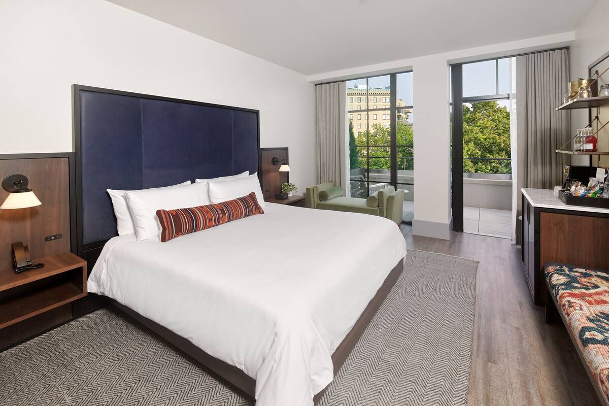 A modern hotel room with a large bed, a desk, a chair, and a view of the city through large windows.