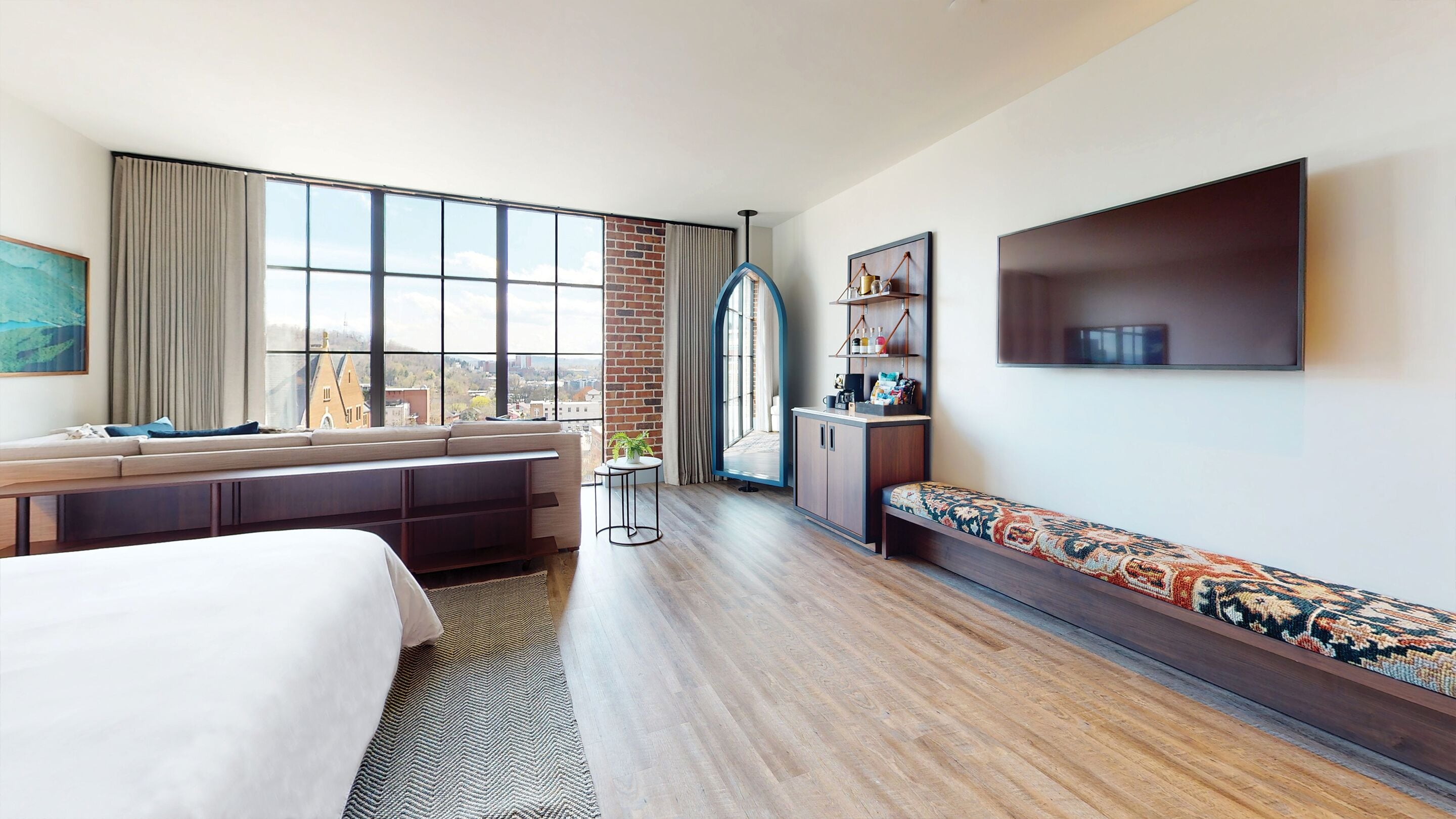 A hotel room with a large window, a sofa, a bed, a TV, and a small table with a plant.