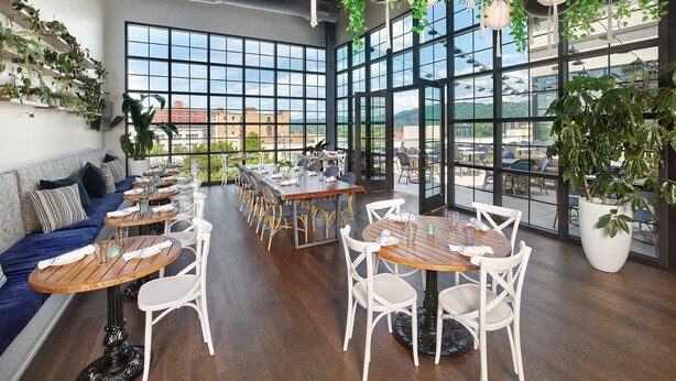 A modern restaurant with large windows, wooden tables, white chairs, and hanging plants. - The Restoration Asheville (Asheville)