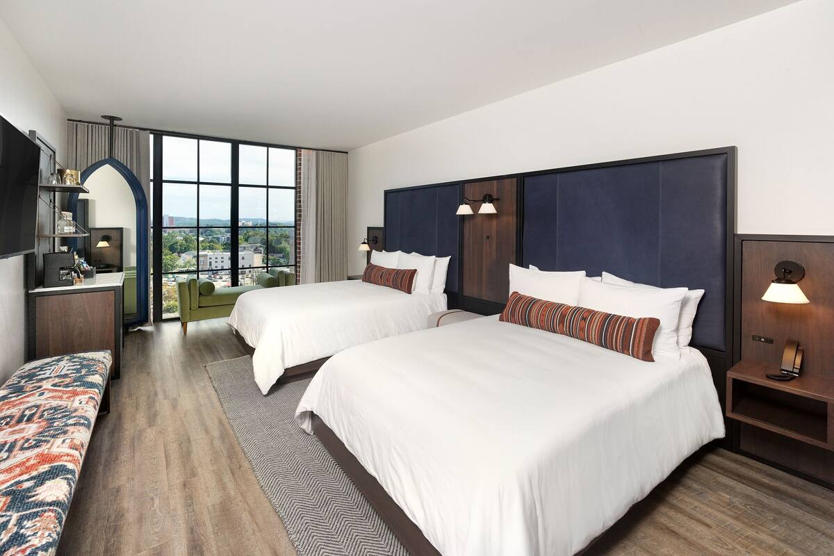 A hotel room with two beds, a desk, a chair, and a view of the city.