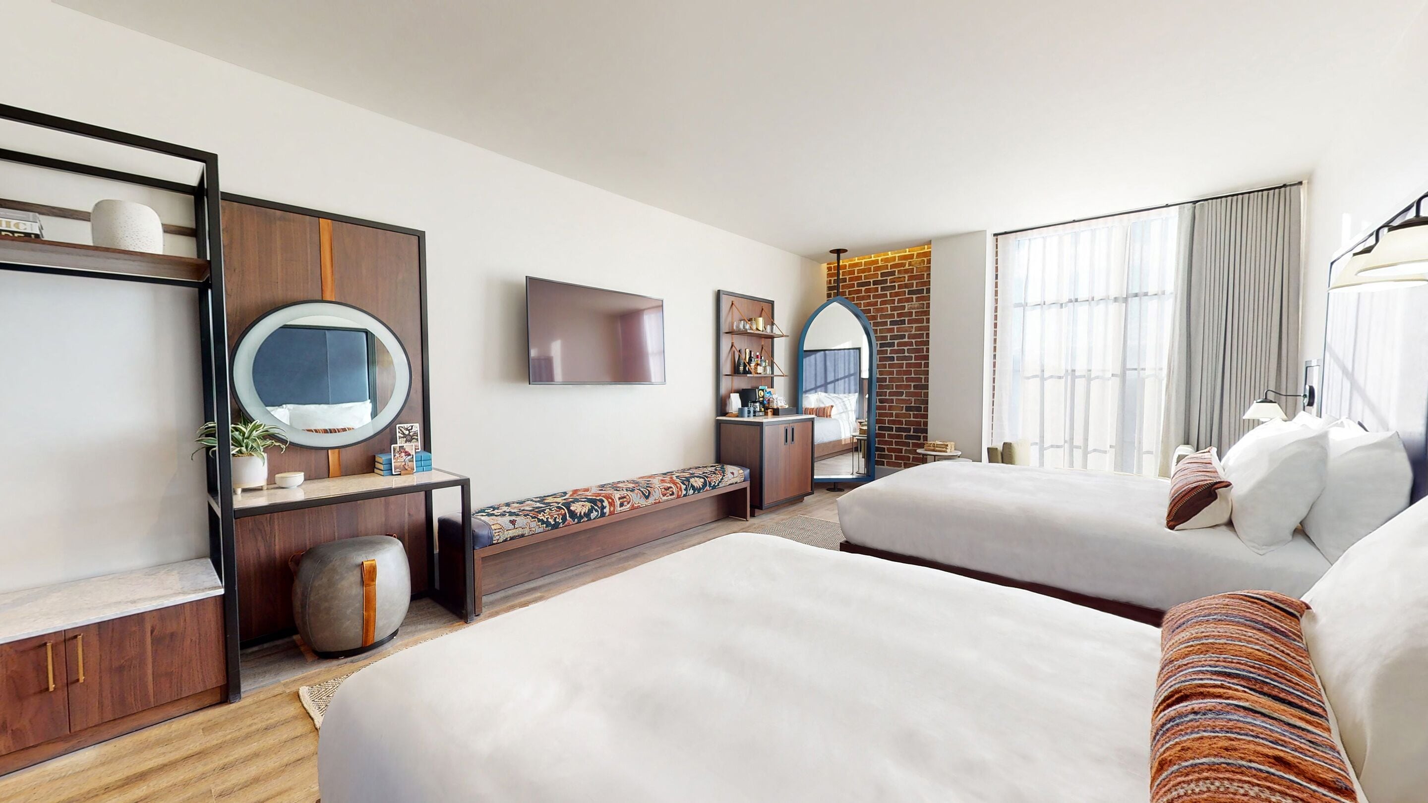 Habitación de hotel con dos camas, televisor, espejo, escritorio pequeño y pared decorativa de ladrillo.