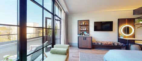 Una habitación de hotel moderna con ventanas amplias, una cama, zona de estar, minibar y televisión.