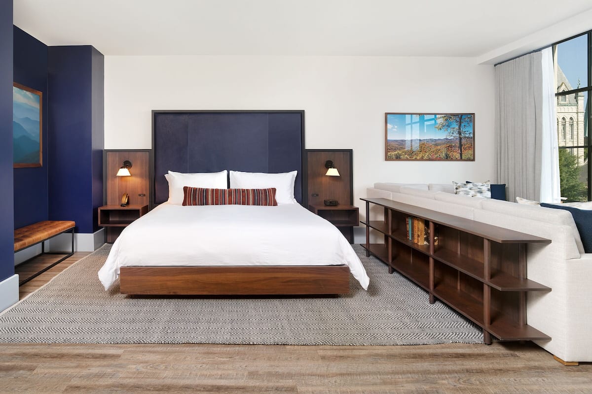 A modern hotel room with a large bed, two bedside tables with lamps, a wooden bench, a sofa, and a view of a building through a window.