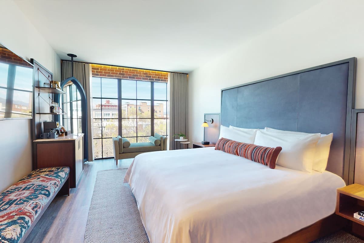 A hotel room with a large bed, a desk with a computer, a bench, and a view of buildings through large windows.