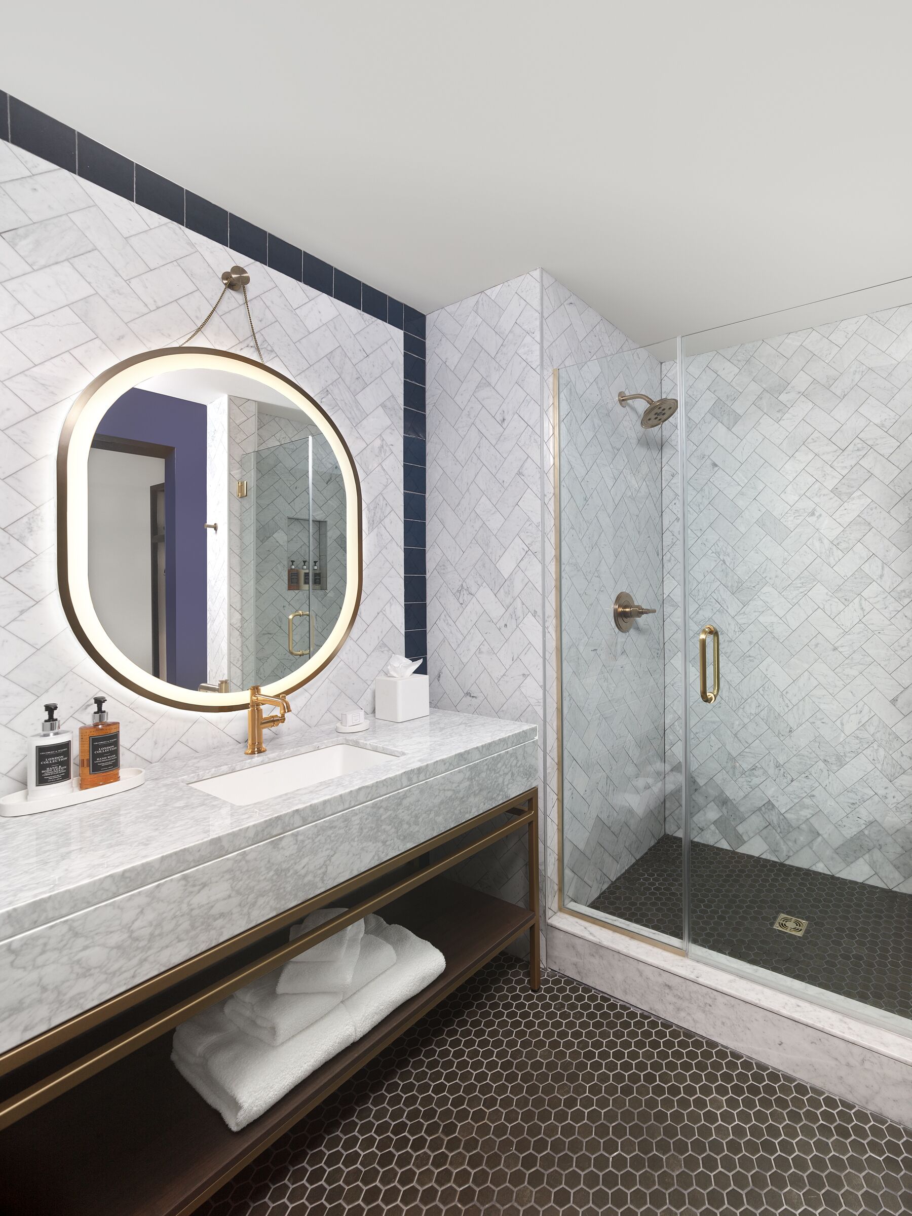 A modern bathroom with a marble vanity, a round mirror, a glass-enclosed shower, and a hexagonal tile floor.