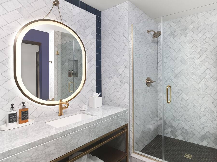 Um banheiro moderno com toucador de mármore, espelho redondo, chuveiro com divisória de vidro e piso de azulejos hexagonais.