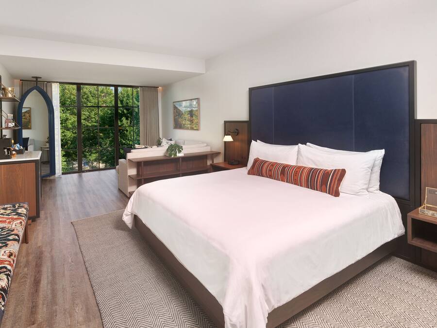 A modern hotel room with a large bed, a desk, and a view of greenery through the window.