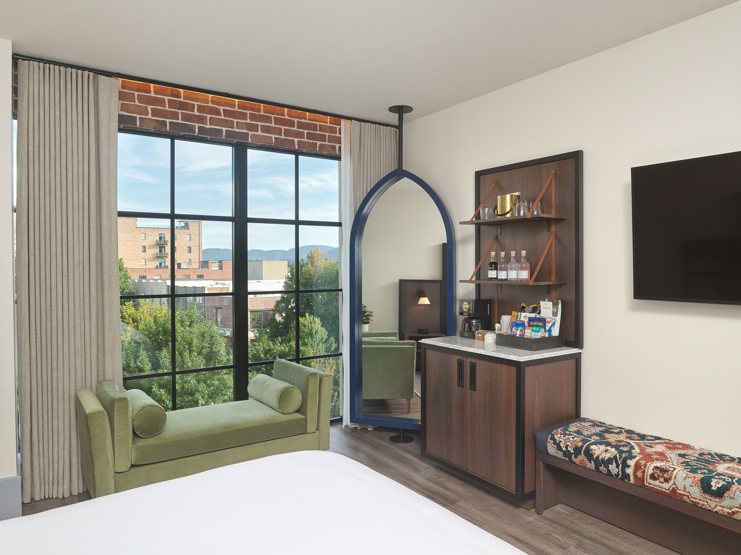 Habitación de hotel con un ventanal grande, un sofá verde, un mueble de madera con estantes y un televisor de pantalla plana en la pared.