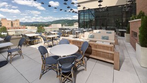 An outdoor patio with round tables and wicker chairs, a wooden bench, and string lights against a backdrop of a modern building and distant mountains. - The Restoration Asheville (Asheville)