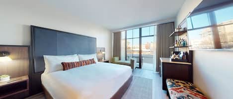 A modern hotel room with a large bed, a bedside table with a lamp, a small seating area, a large window with a view of the city, and a minibar.