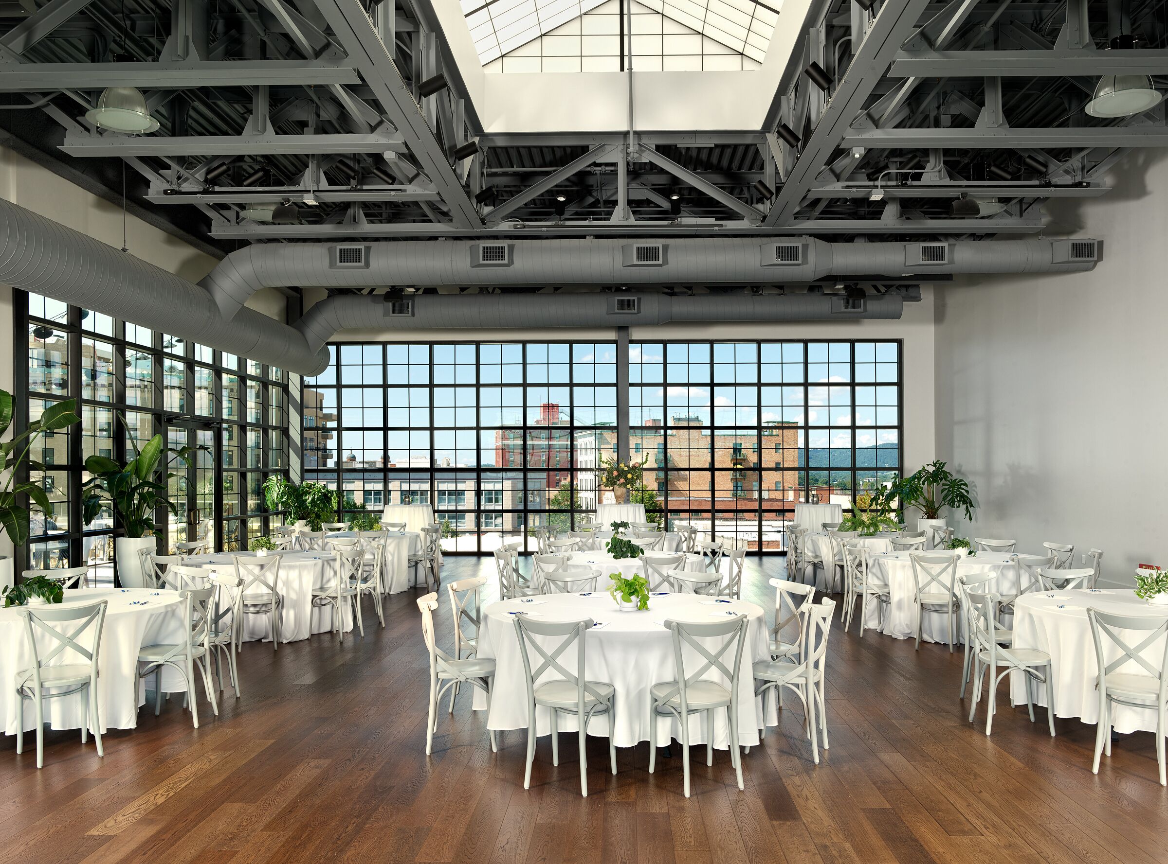 A spacious indoor dining area with multiple tables and chairs, large windows offering a city view, and a high ceiling with visible structural beams.