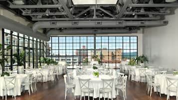 A spacious indoor dining area with multiple tables and chairs, large windows offering a city view, and a high ceiling with visible structural beams.