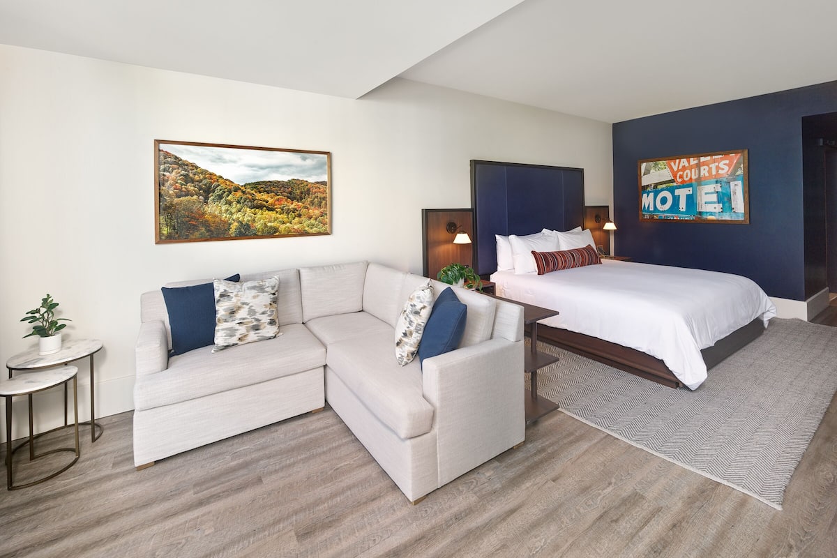 A modern hotel room with a sectional sofa, a single bed, a small table with a plant, and a large landscape painting on the wall.