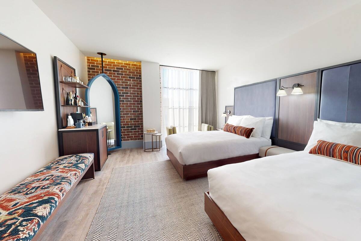 A hotel room with two beds, a desk, a mirror, a brick accent wall, and a large window.