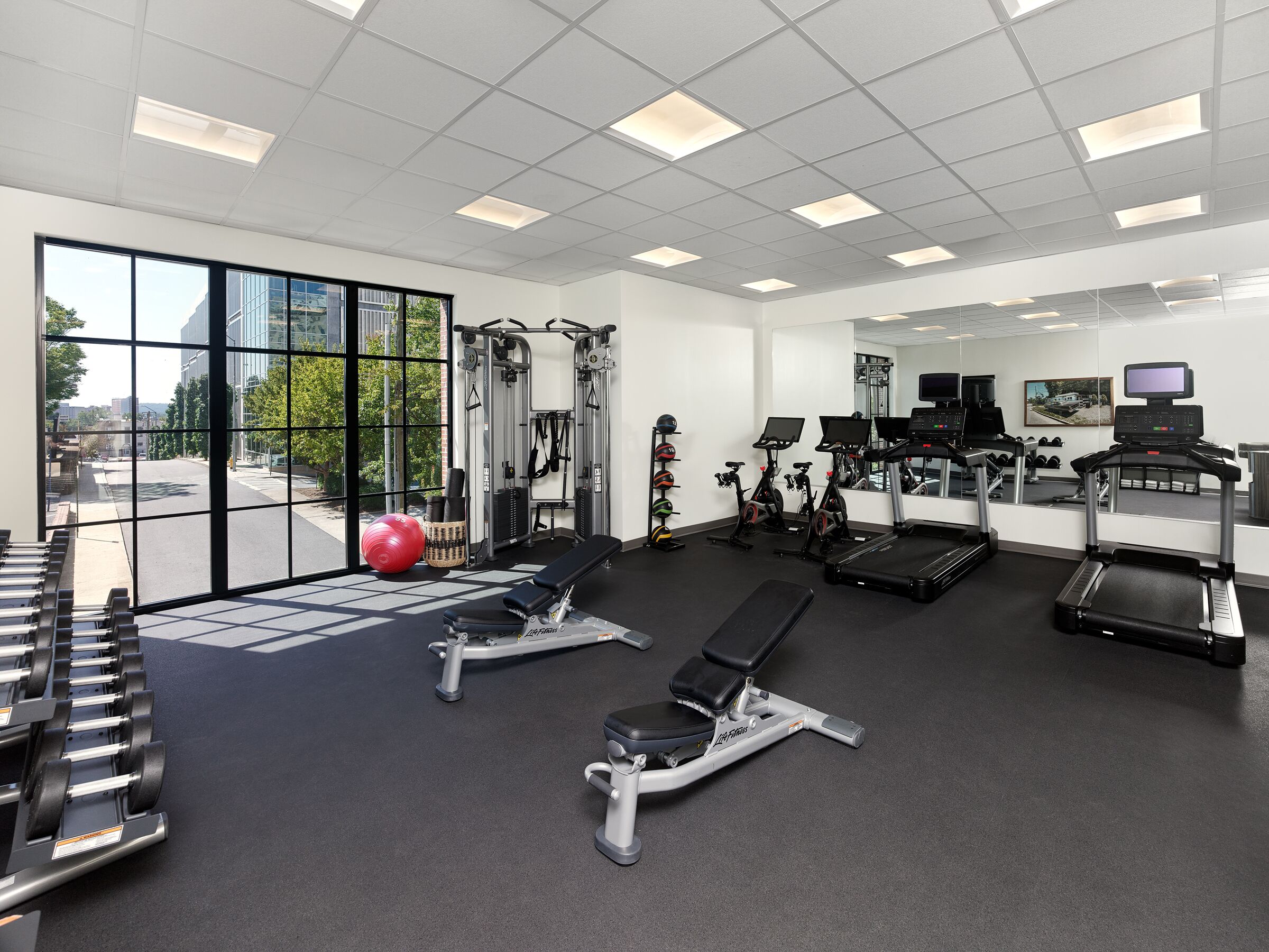 A gym with exercise equipment, a bench, a treadmill, and a large window with a view of a building and trees.
