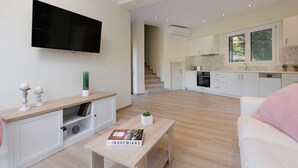 En moderne stue med et fladskærms TV, et træ sofabord og en sofa. Et køkken med hvide skabe og et vindue kan ses i baggrunden.