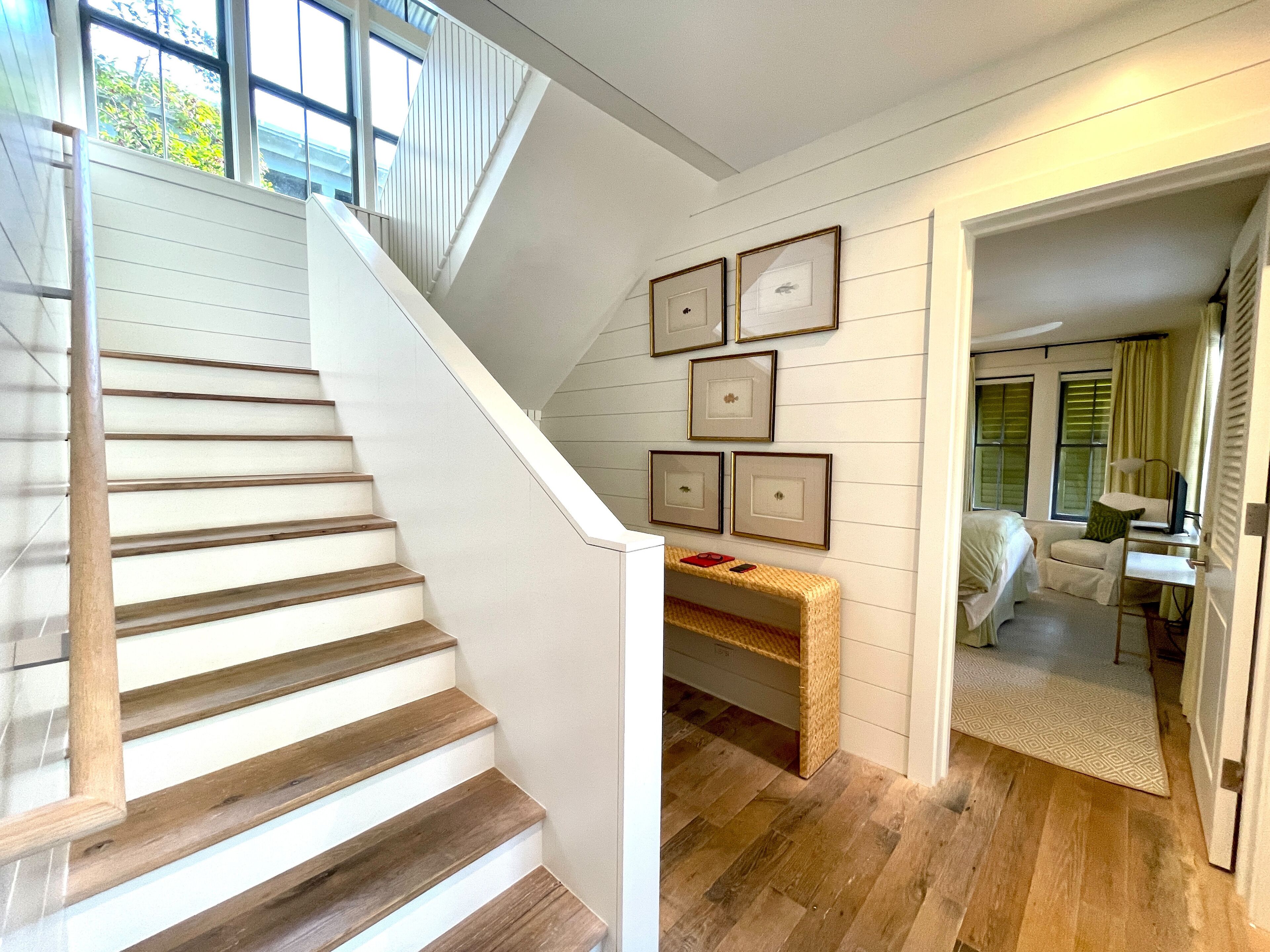 A staircase with wooden steps and white walls, leading to a room with a desk, chair, and a bed.