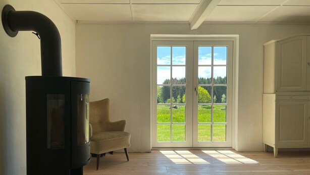 Een kamer met een zwart houtkachel, een beige fauteuil en uitzicht door een glazen deur op een groen landschap.
