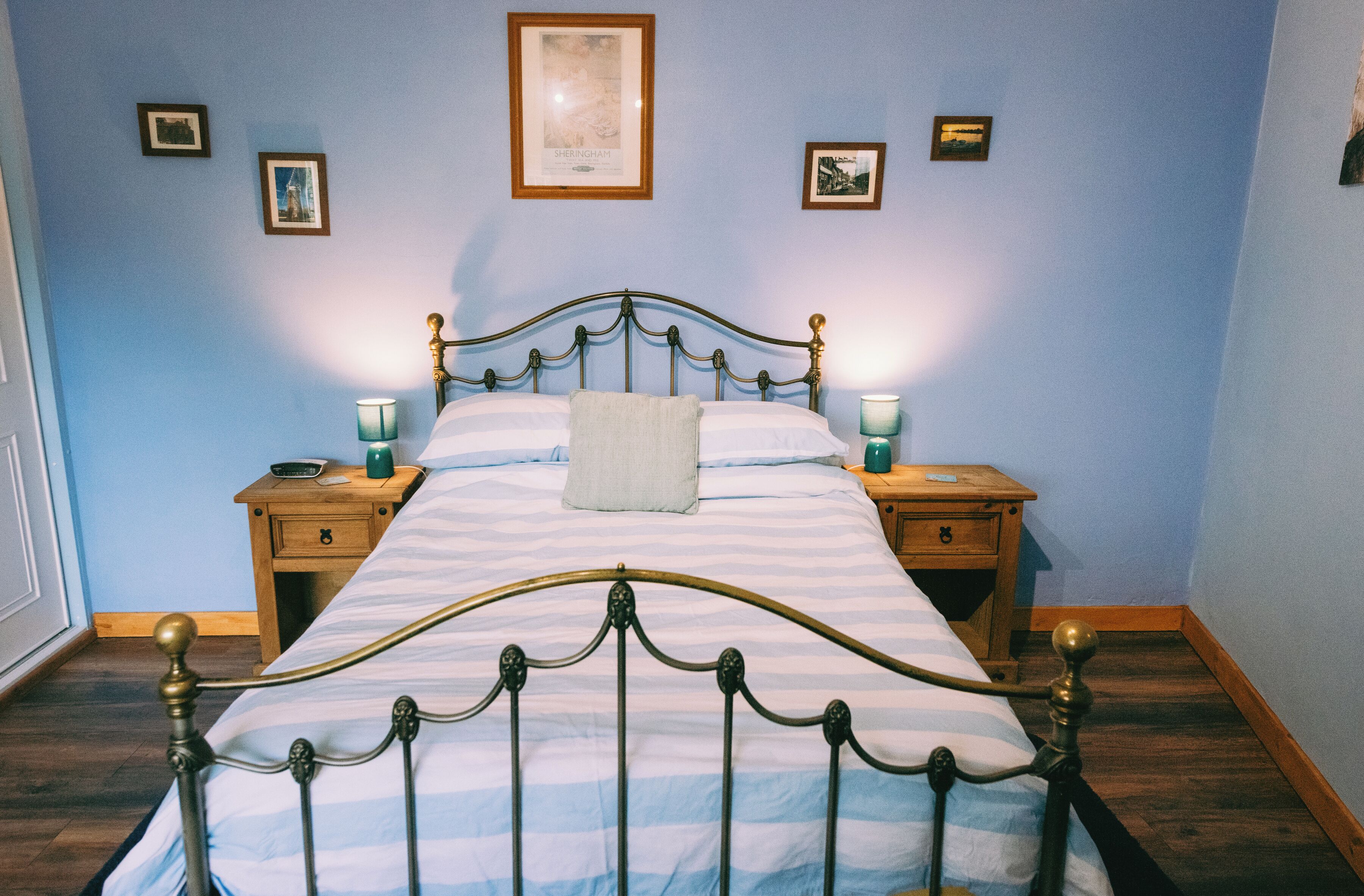 Una cama bien tendida con una colcha a rayas y una sola almohada, flanqueada por dos mesitas de noche con lámparas, en una habitación con paredes azul claro y cuadros enmarcados.