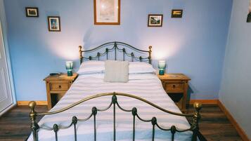 Una cama bien tendida con una colcha a rayas y una sola almohada, flanqueada por dos mesitas de noche con lámparas, en una habitación con paredes azul claro y cuadros enmarcados.
