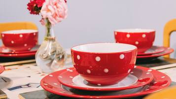 Una mesa con tazas y platillos rojos y blancos con lunares, un florero transparente con flores rosas, y una silla amarilla al fondo.