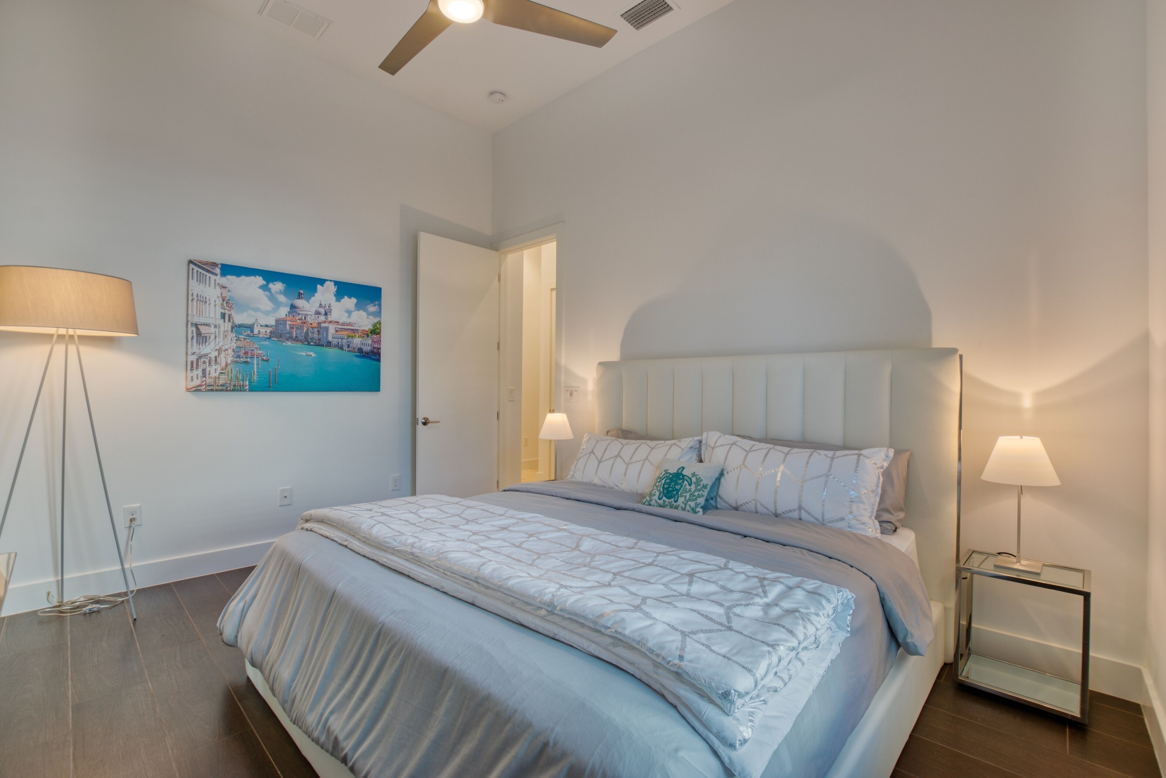 Une chambre à coucher avec un lit, une table de chevet, une lampe, un tableau et un ventilateur de plafond.
