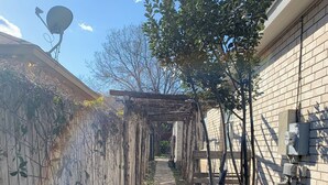 A narrow stone pathway flanked by a wooden fence and a brick wall, leading to a covered area with a pergola.