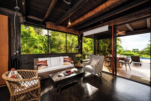 Un salon moderne avec un canapé, un fauteuil et une table basse, de grandes fenêtres offrant une vue sur une végétation luxuriante, et un espace piscine visible à travers une porte ouverte.