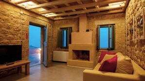 A cozy living room with a stone wall, a fireplace, a television, and a sofa with cushions. - Patio Villas (West Mani)