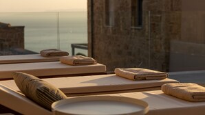 A sunlit room with two sunbeds, each with a towel and pillow. A stone wall and a window with a view of the sea are visible. - Patio Villas (West Mani)