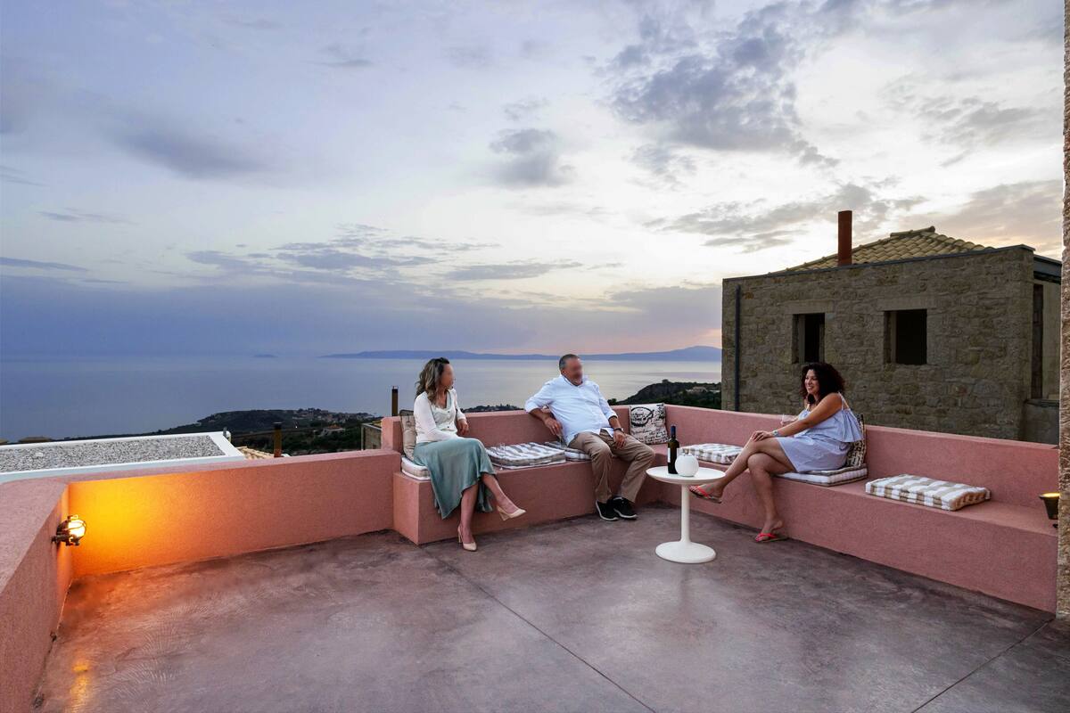 A rooftop terrace with a stone building, seating areas, and a view of the sea.
