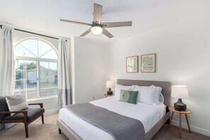 A hotel room with a bed, two bedside tables with lamps, a chair, a ceiling fan, a large window with curtains, and two wall art pieces.