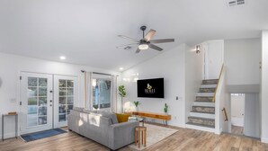 A modern living room with a grey sofa, wooden flooring, a ceiling fan, a TV mounted on the wall, and a staircase with patterned tiles. - Mesa Getaway | Htd. Pool, Pellet Grill & Patio TV (Mesa)