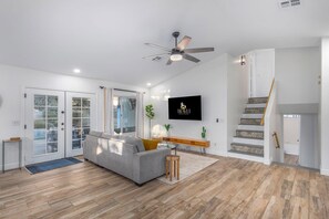 A modern living room with a grey sofa, wooden flooring, a ceiling fan, a TV mounted on the wall, and a staircase with patterned tiles.