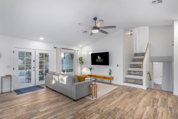 A modern living room with a grey sofa, wooden flooring, a ceiling fan, a TV mounted on the wall, and a staircase with patterned tiles.