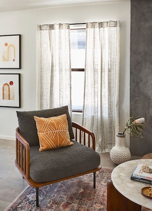 A modern living room with a dark grey armchair, a patterned rug, a round coffee table, and a window with sheer curtains.